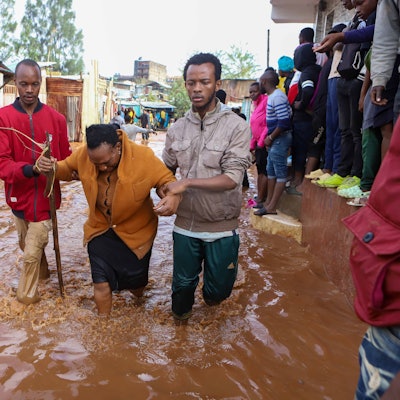Eine Frau mit einem Gehstock wird von zwei Männern gestützt. Sie stehen bis zu den Knien in bräunlichem Wasser.
