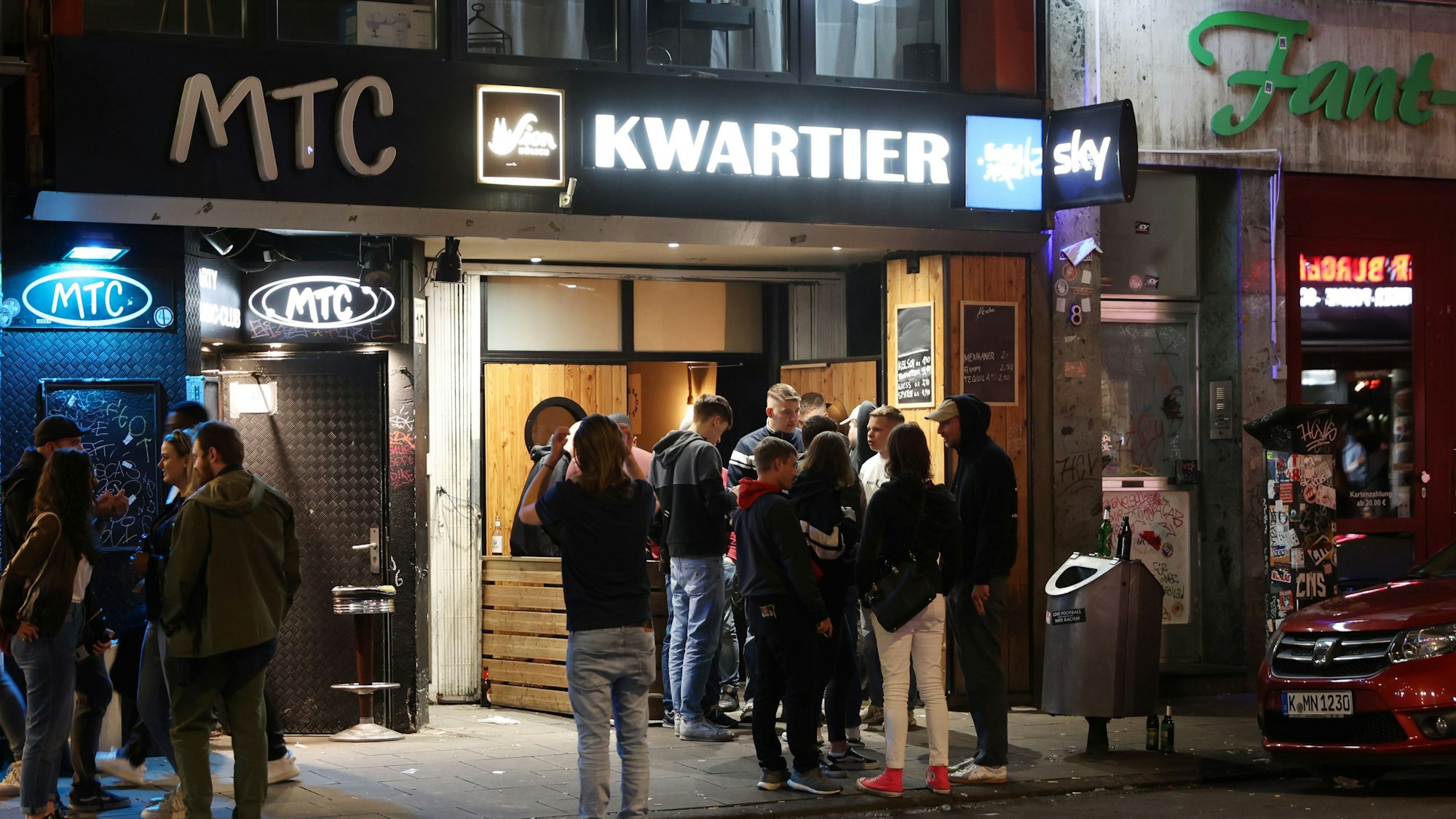 Fassade mit Bars und Clubs, davor stehen trinkende Menschen.