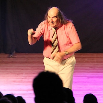 Guildo Horn steht auf der Bühne des Euskirchener Stadttheaters. Er trägt ein lachsfarbenes, kurzärmeliges Hemd mit einer braun-gestreiften Krawatte und eine helle Hose.