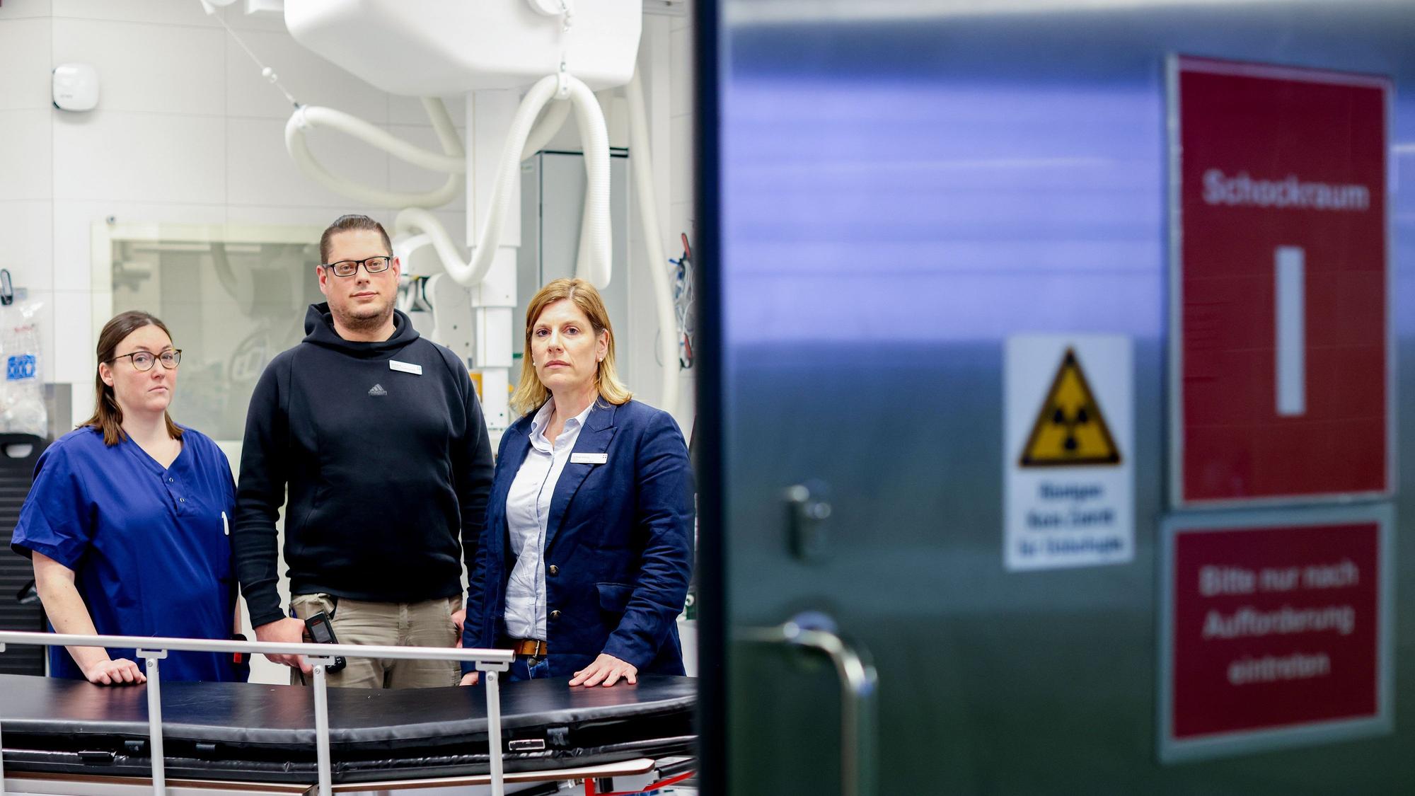 Drei Menschen stehen in der Notaufnahme eines Krankenhauses und schauen in die Kamera.
