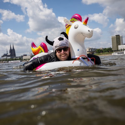 Ein kostümierter Teilnehmer schwimmt im Rhein am Panorama der Stadt mit dem Kölner Dom vorbei.