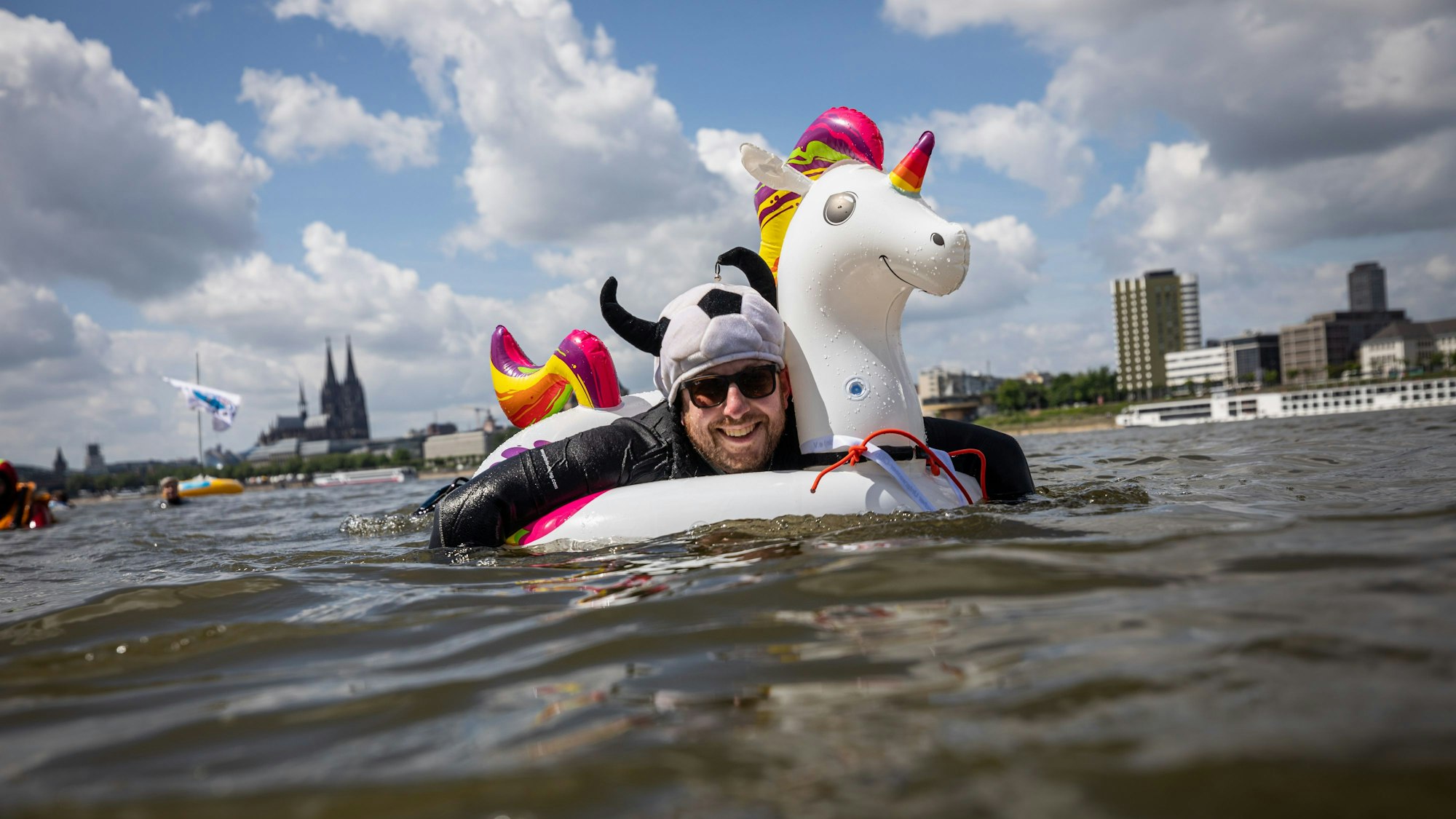 Ein kostümierter Teilnehmer schwimmt im Rhein am Panorama der Stadt mit dem Kölner Dom vorbei.