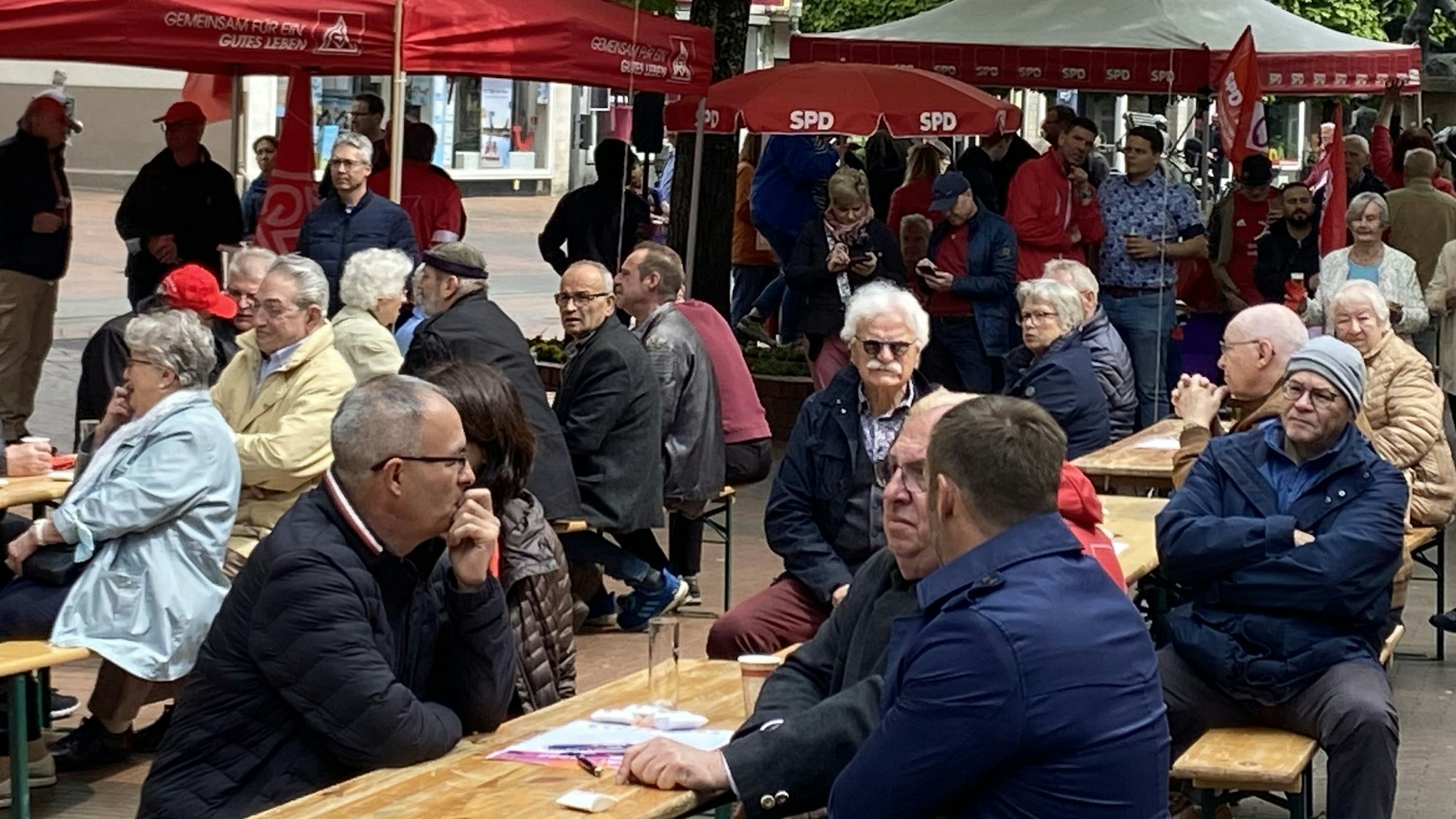 Das Foto zeigt die Mai-Kundgebung aus 2022. Frauen und Männer sitzen an Biertischen und hören den Rednerinnen und Rednern zu.