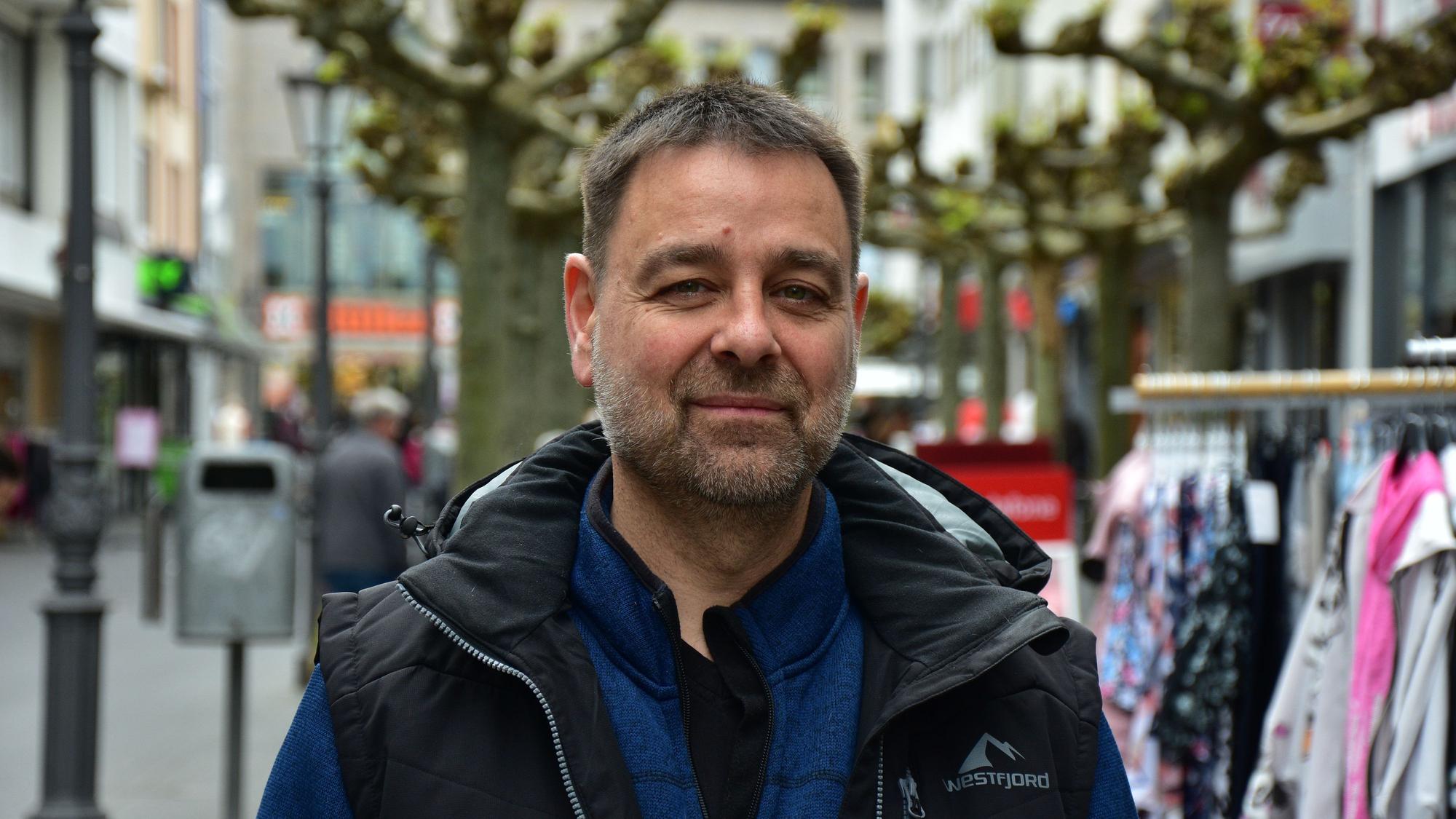 Ein Mann mit Bart in einer Fußgängerzone. Er trägt eine blaue Jacke und darüber eine schwarze Weste. im Hintergrund stehen Platanen und Ständer mit Kleidung vor Geschäften.