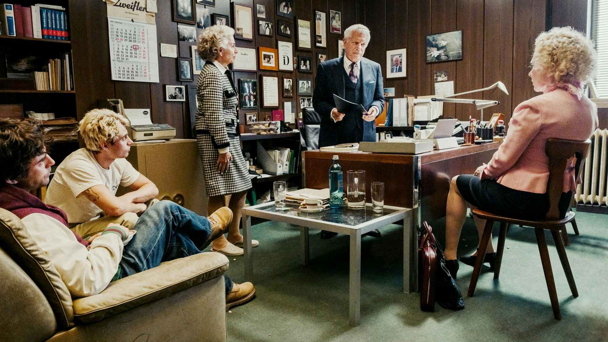 Symcha Zweifler steht hinter seinem Schreibtisch. Er schaut ernst, hält eine Mappe in seiner Hand. Seine Frau Lilka steht an der Ecke des Tisches, seine Tochter Mimi sitzt auf dem Stuhl ihm gegenüber. Die Enkel Samuel und Leon sitzen auf einer Couch an der Seite. Alle stehen Symcha ernst an. Im Hintergrund hängen Urkunden, Fotos, Kalender und andere Erinnerungen an der Wand.