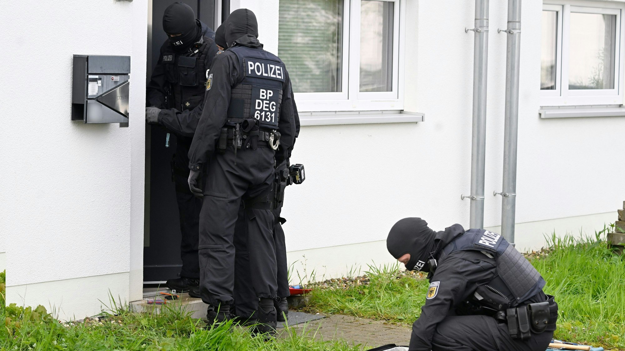 Ein vermummter Beamter kniet auf dem Boden, zwei gehen in eine geöffnete Haustür: Polizisten durchsuchen am 18. April Wohnungen und Häuser in Düren bei der Razzia gegen Schleuserkriminalität.