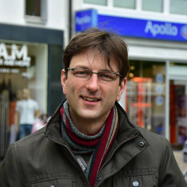 Ein Mann mit Brille, Schal und brauner Winterjacke vor Geschäften in einer Fußgängerzone.