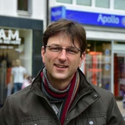 Ein Mann mit Brille, Schal und brauner Winterjacke vor Geschäften in einer Fußgängerzone.