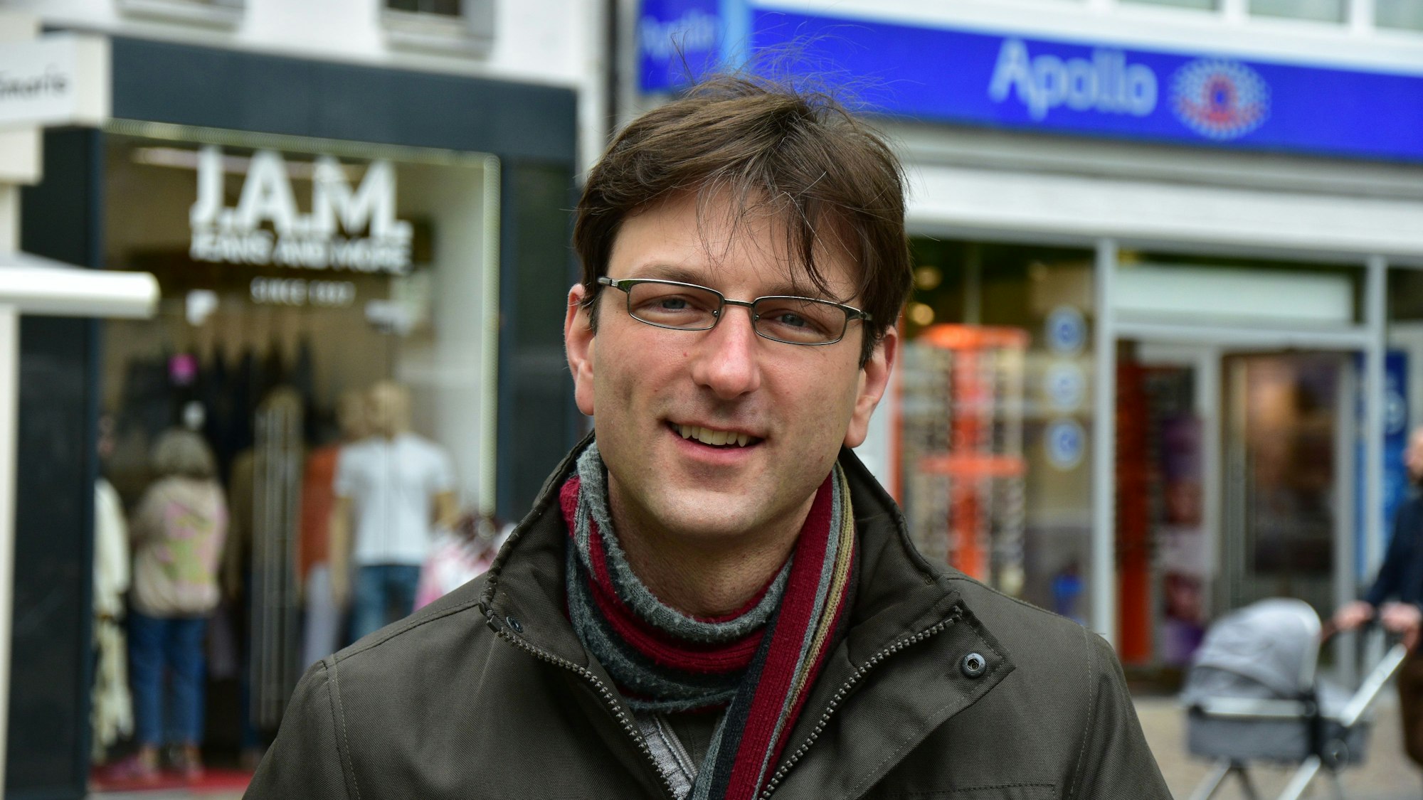 Ein Mann mit Brille, Schal und brauner Winterjacke vor Geschäften in einer Fußgängerzone.