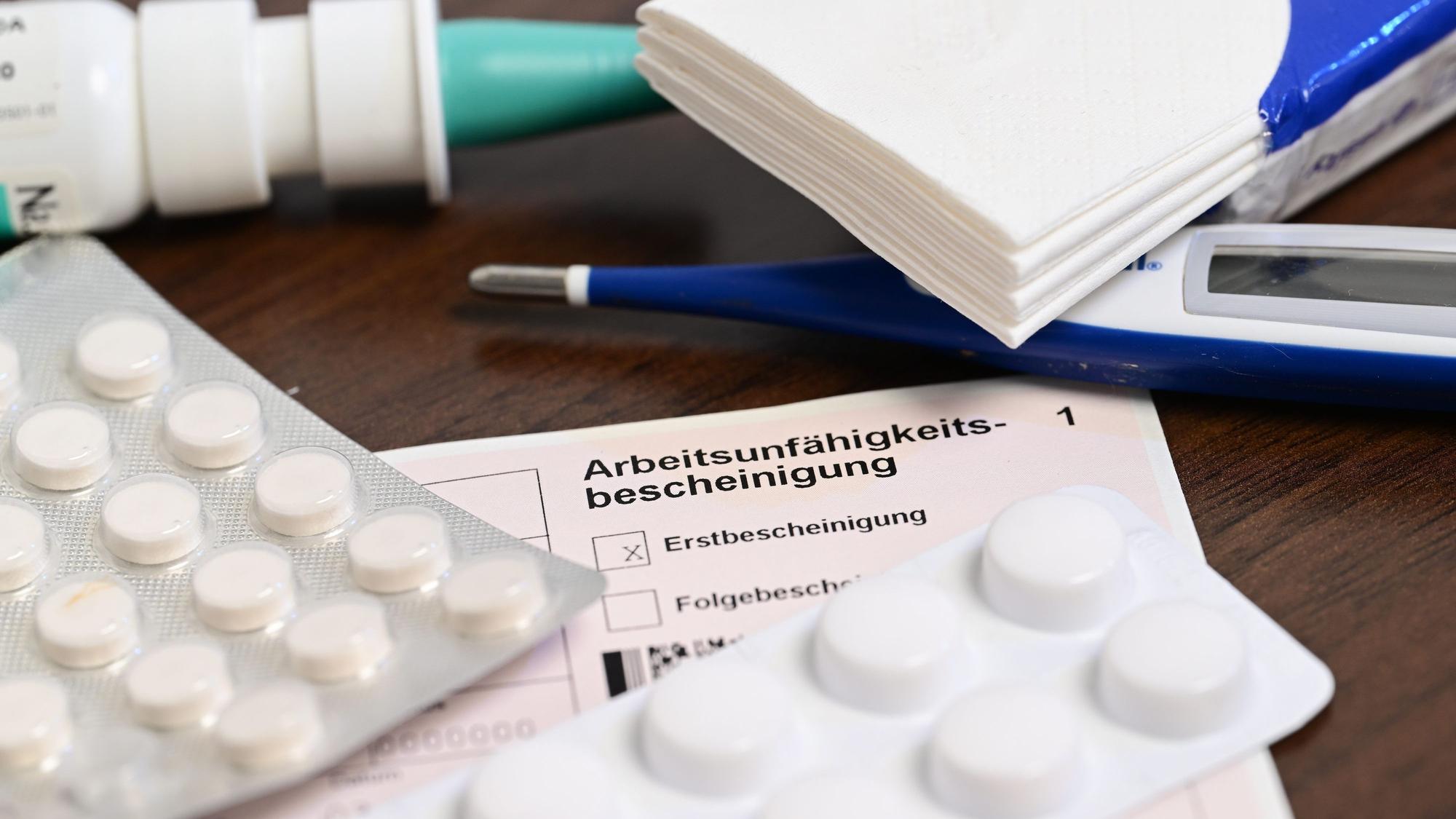 Ein Fieberthermometer, Medikamente und eine ärztliche Arbeitsunfähigkeitsbescheinigung (Krankmeldung) liegen auf einem Nachttisch.