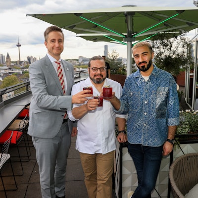 Drei Männer stehen mit Drinks in der Hand auf der Terrasse einer Rooftop-Bar.