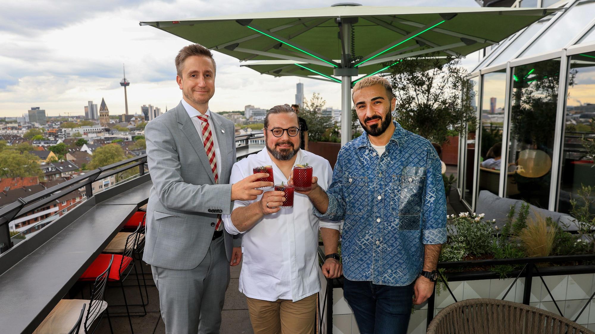 Drei Männer stehen mit Drinks in der Hand auf der Terrasse einer Rooftop-Bar.