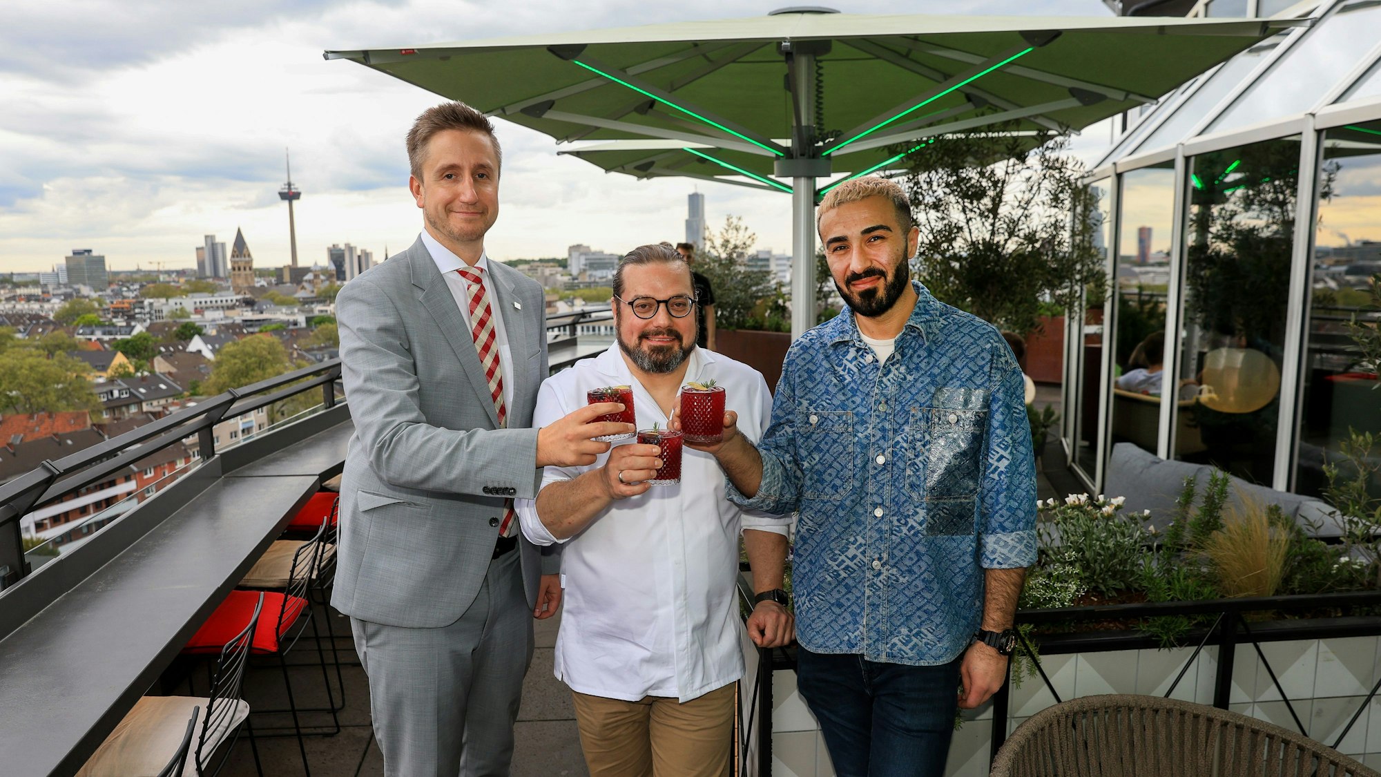 Drei Männer stehen mit Drinks in der Hand auf der Terrasse einer Rooftop-Bar.
