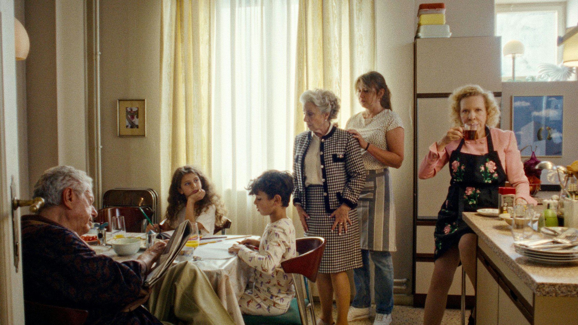 Großvater Symcha sitzt im linken Bildrand am Esstisch und liest Zeitung, die Zwillinge machen Hausaufgaben am Tisch. Gromutter Lilka schaut auf sie herab, hinter die Haushälterin, sie auf sie achtet. Mimi Zweifler sitzt an der Küchenecke und trinkt einen Kaffee.