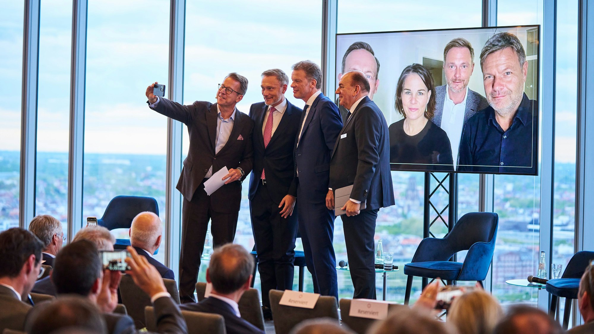Jubiläumsfeier Flossbach von Storch. Moderator Ulrich Stockheim nutzt die Chance für ein Foto mit Christian Lindner, Christian Sewing und Axel Weber (v.l). Auf dem Bildschirm im Hintergrund ist Lindner mit einigen seiner Kabinettskollegen beim Selfie am Tag der Bildung der Ampelkoalition zu sehen.