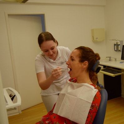 Celine Doerr blickt in der Zahnarztpraxis, in der sie arbeitet, in den Mund einer Patientin, die auf dem Behandlungsstuhl sitzt.