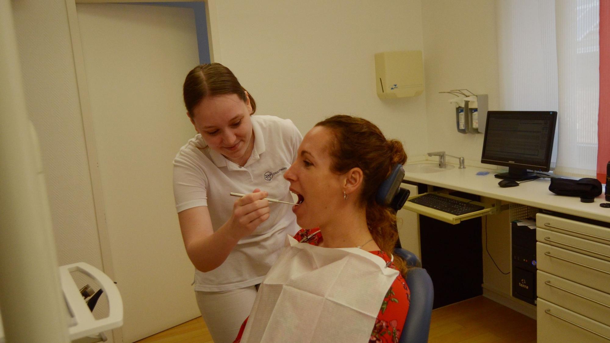 Celine Doerr blickt in der Zahnarztpraxis, in der sie arbeitet, in den Mund einer Patientin, die auf dem Behandlungsstuhl sitzt.