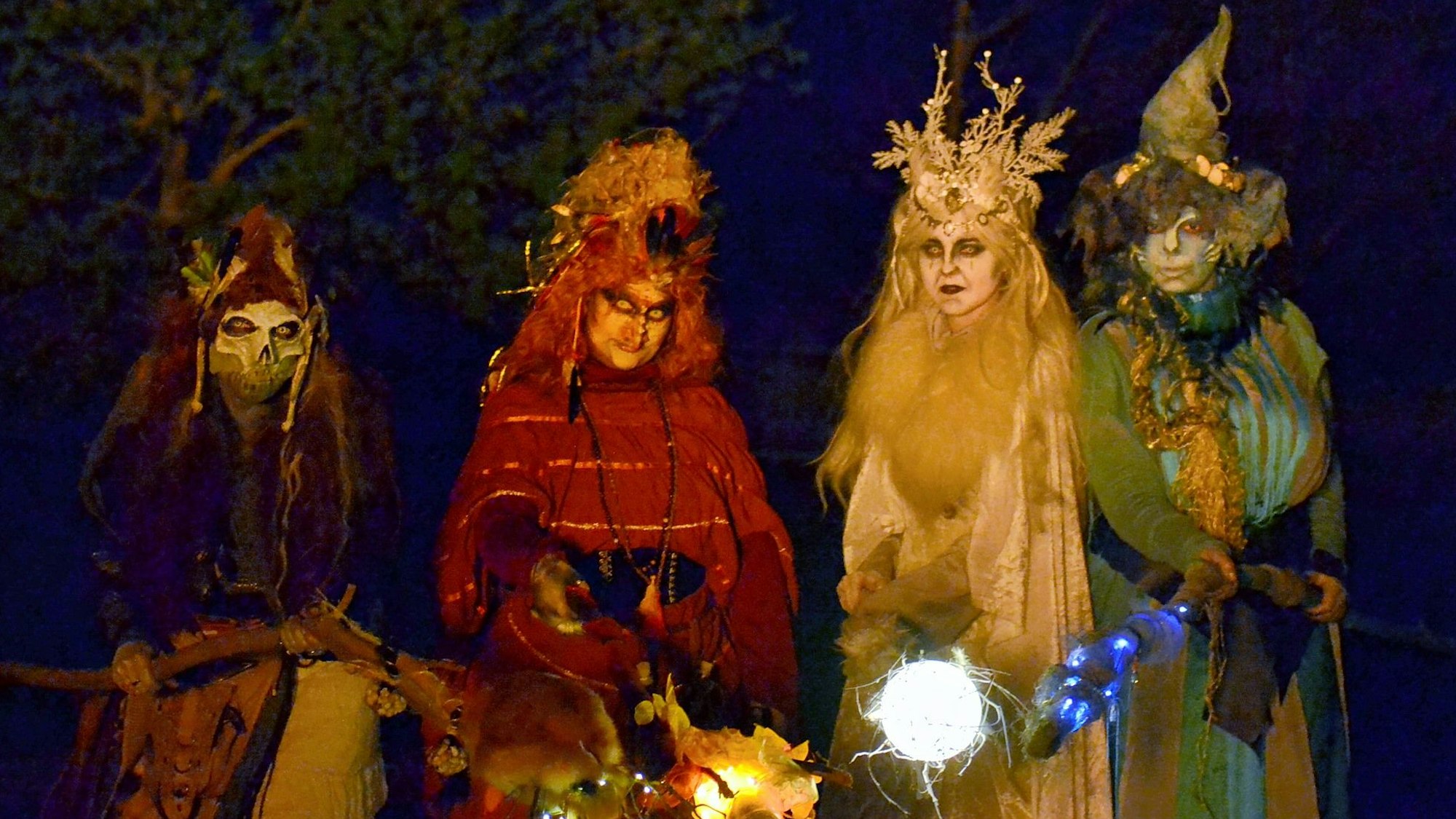 Vier farbenfroh kostümierte Hexen mit leuchtenden Fackeln in der Dunkelheit.