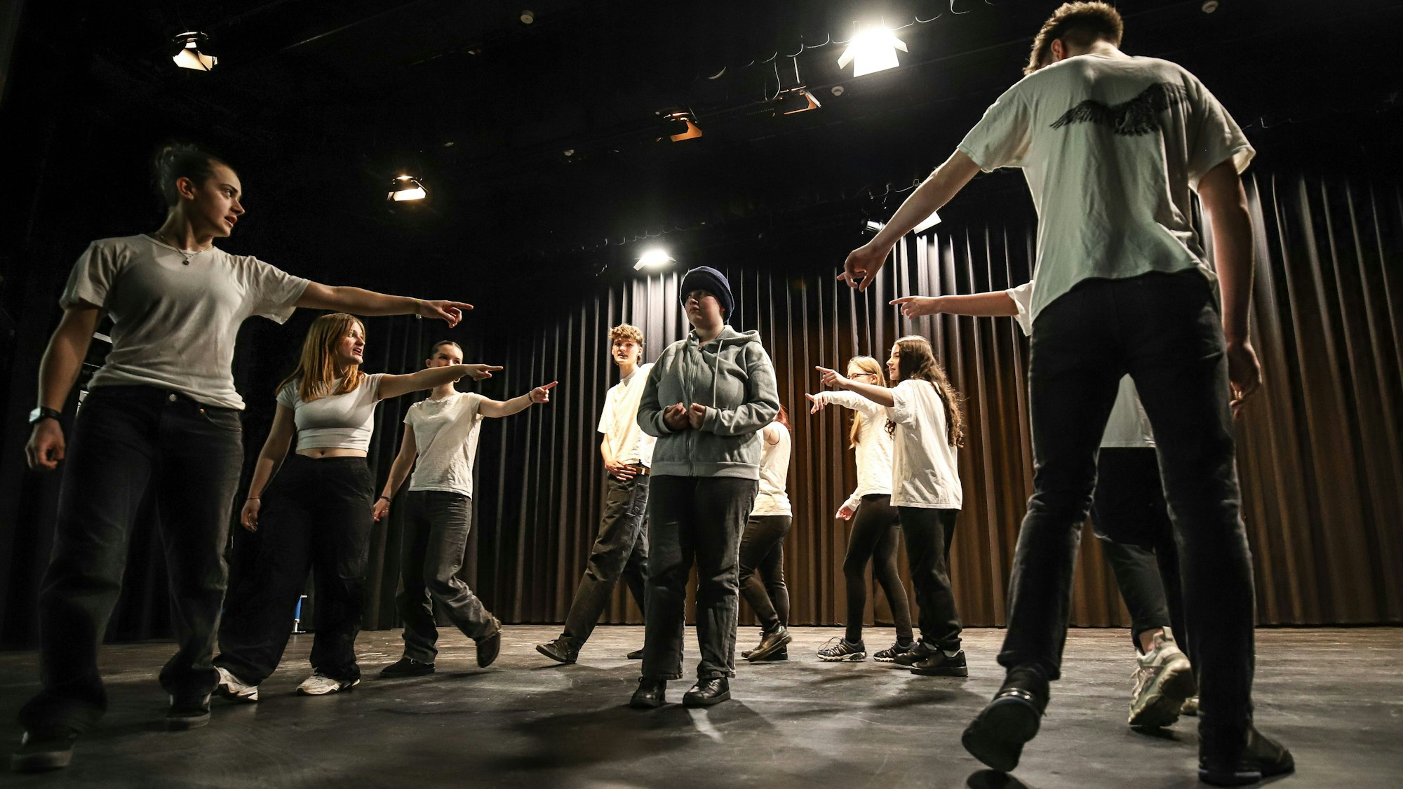 Schülerinnen und Schüler stehen im Kreis auf einer Bühne und zeigen mit dem Finger auf einen Jungen.