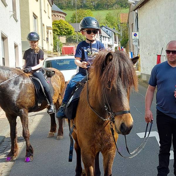 Zwei junge Teilnehmer reiten am Aachener Bischof in seinem roten Ornat vorbei. Eines der Pferde wird von einem Helfer geführt.