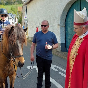 Zwei junge Teilnehmer reiten am Aachener Bischof in seinem roten Ornat vorbei. Eines der Pferde wird von einem Helfer geführt.
