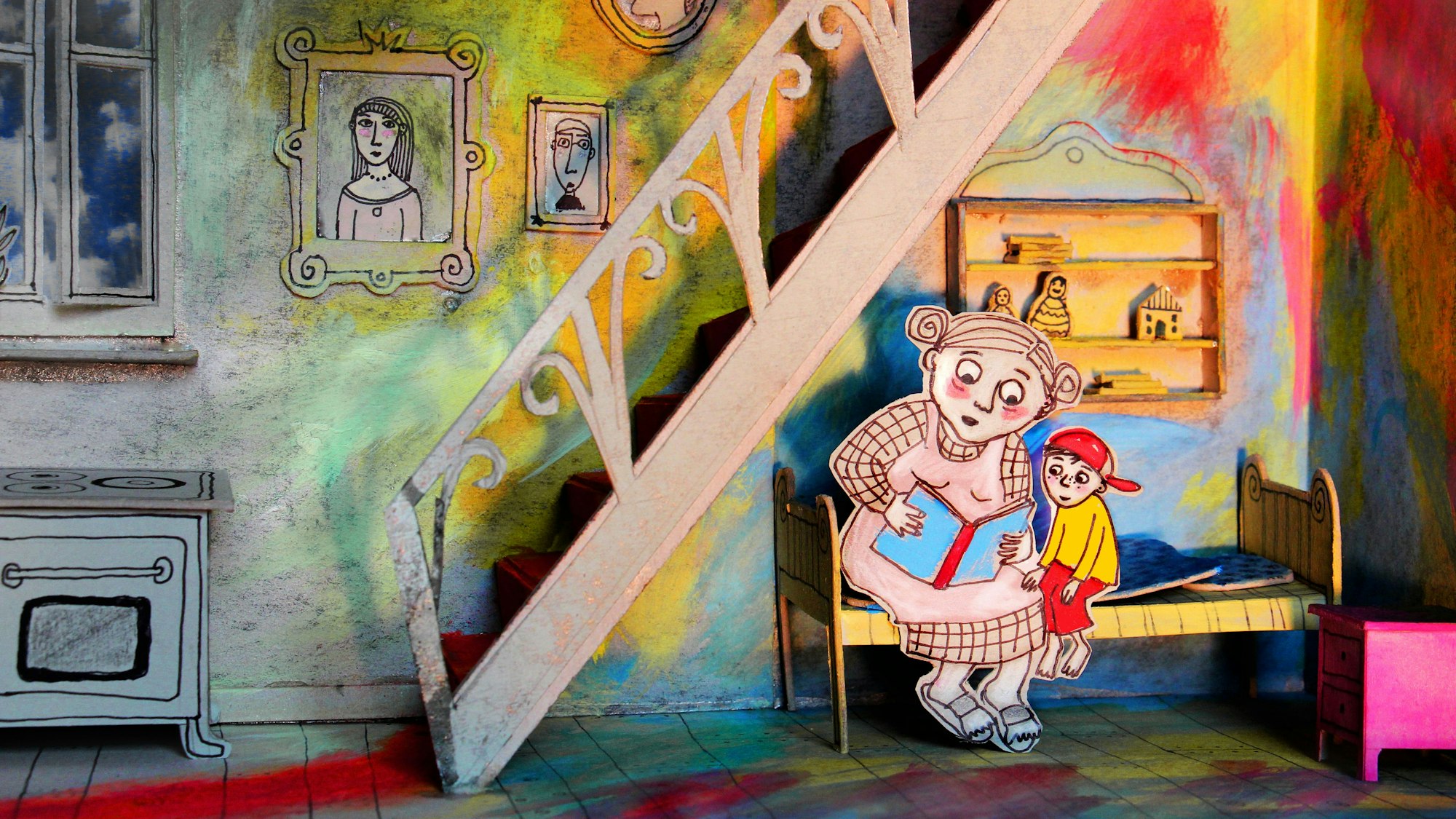 Eine Bilderbuchszene in einer Art kleiner Bühne. Eine aus Papier augeschnittene Frau sitzt mit einem kleinen Jungen auf dem Bett und liest ihm vor. Die Wände und Möbel sind in leuchtenden Farben gestaltet.