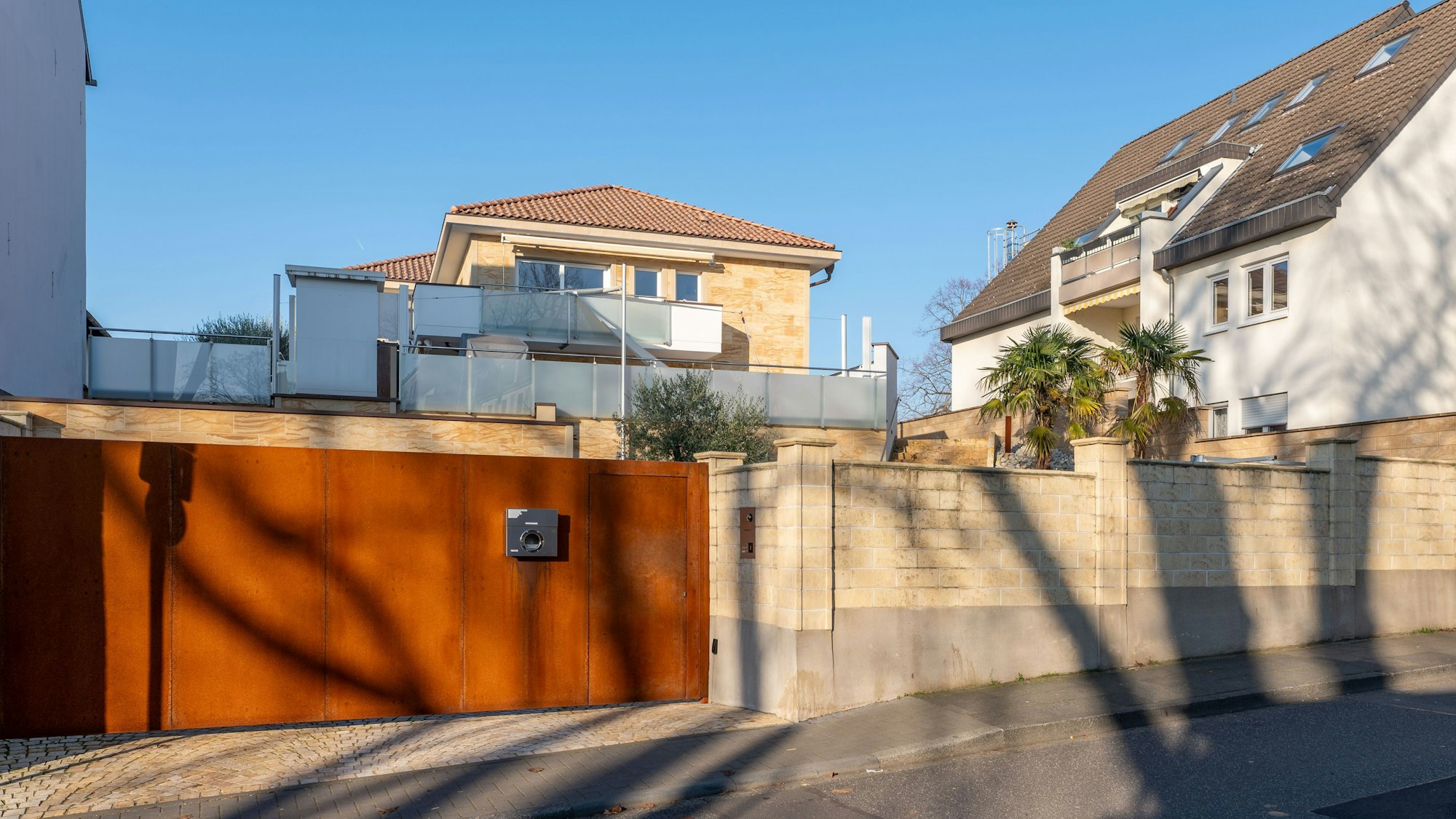 Außenansicht auf die Immobilie in Niederkassel-Mondorf: das Grundstück ist über ein rostfarbenes massives Eingangstor zu erreichen. Dahinter ist die sandsteinfarbene Villa zu sehen.