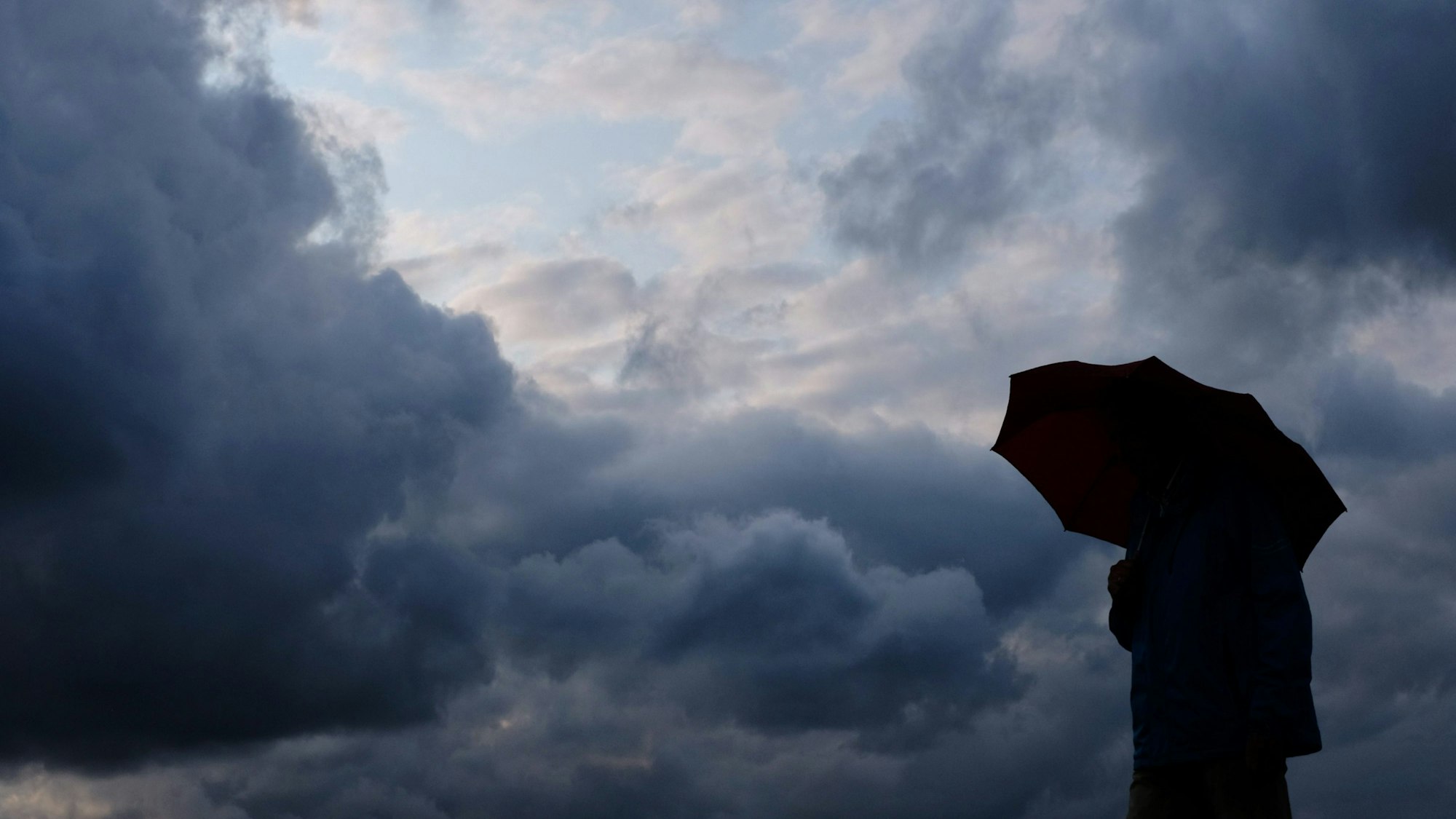 Ein Mann geht mit einem Regenschirm vor aufziehenden dunklen Wolken spazieren. (Symbolfoto)