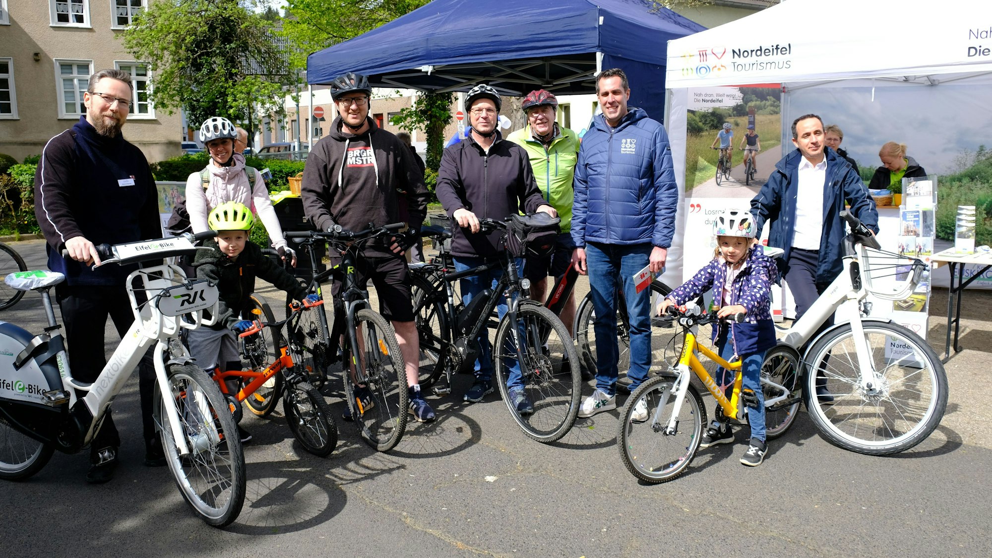 Mehrere Radfahrer, darunter zwei Kinder, stehen mit ihren Fahrrädern in Schleiden vor zwei Zelt-Pavillons.