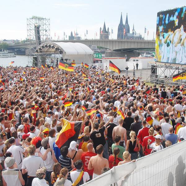 Eine Großleinwand steht auf der Deutzer Werft, im Hintergrund ist der Kölner Dom zu sehen. Auf der Fläche davor stehen viele hundert Fußballfans.