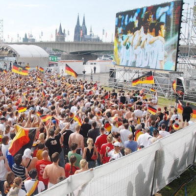 Eine Großleinwand steht auf der Deutzer Werft, im Hintergrund ist der Kölner Dom zu sehen. Auf der Fläche davor stehen viele hundert Fußballfans.