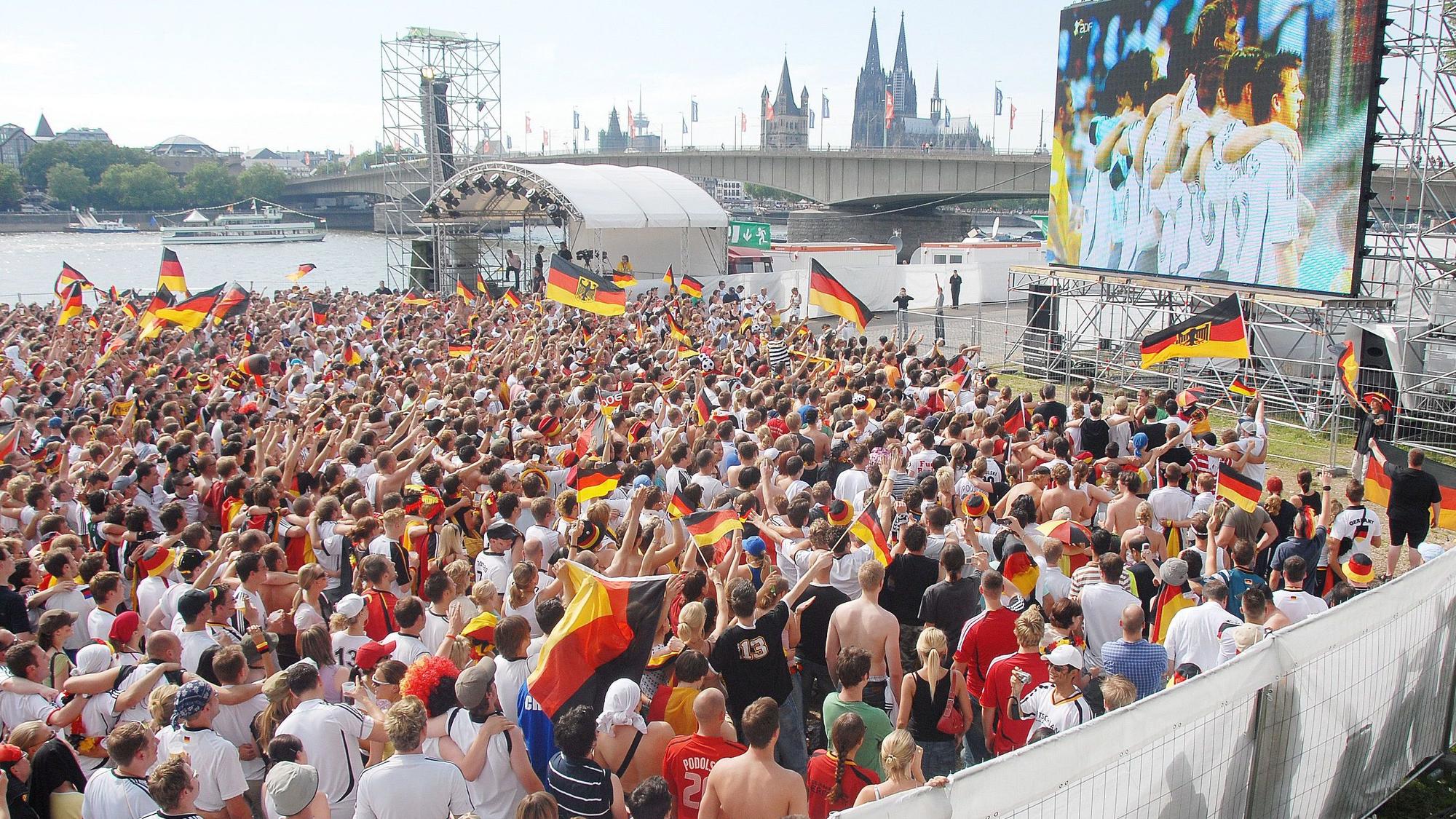Eine Großleinwand steht auf der Deutzer Werft, im Hintergrund ist der Kölner Dom zu sehen. Auf der Fläche davor stehen viele hundert Fußballfans.