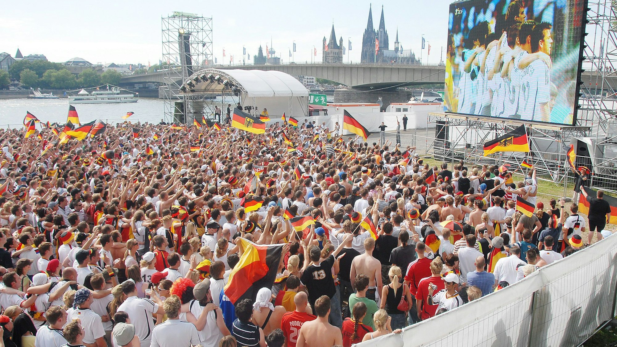 Eine Großleinwand steht auf der Deutzer Werft, im Hintergrund ist der Kölner Dom zu sehen. Auf der Fläche davor stehen viele hundert Fußballfans.