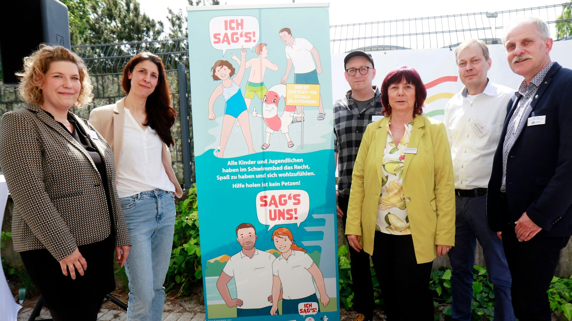 Vertreterinnen und Vertreter des Kölner Netzwerks gegen sexuelle Belästigung in Schwimmbädern stehen zum Start ihrer Kampagne „Ich sag's“ um einen Flyer herum, der ein Kind im Badeanzug zeigt, im Hintergrund ermahnt ein Schwimmmeister einen Jungen, daneben steht ein Rettungsring mit Armen und Beinen, der ein Schild in der Hand hält mit der Aufschrift: Hier ist kein Platz für sexuelle Belästigung.