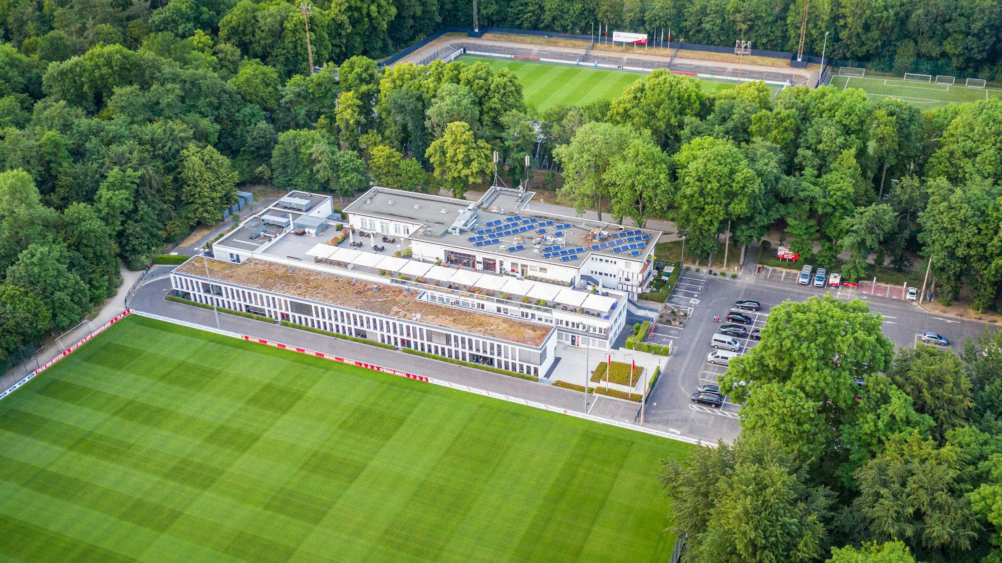 Das Geißbockheim genannte Clubhaus des Fußballvereins 1. FC Köln im Äußeren Grüngürtel mit einem Rasen-Trainingsplatz im Vordergrund und dem Franz-Kremer-Stadion im Hintergrund. Zu sehen auf einer Luftaufnahme, die mithilfe einer Drohne hergestellt wurde.