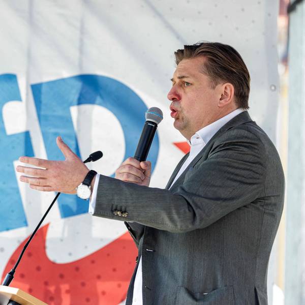 Maximilian Krah auf einer Bühne der Kundgebung in Dresden. Er hält ein Mikrofon an seinen Mund und gestikuliert mit der linken Hand. Im Hintergrund eine Flagge mit AFD-Logo.