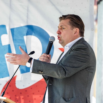 Maximilian Krah auf einer Bühne der Kundgebung in Dresden. Er hält ein Mikrofon an seinen Mund und gestikuliert mit der linken Hand. Im Hintergrund eine Flagge mit AFD-Logo.