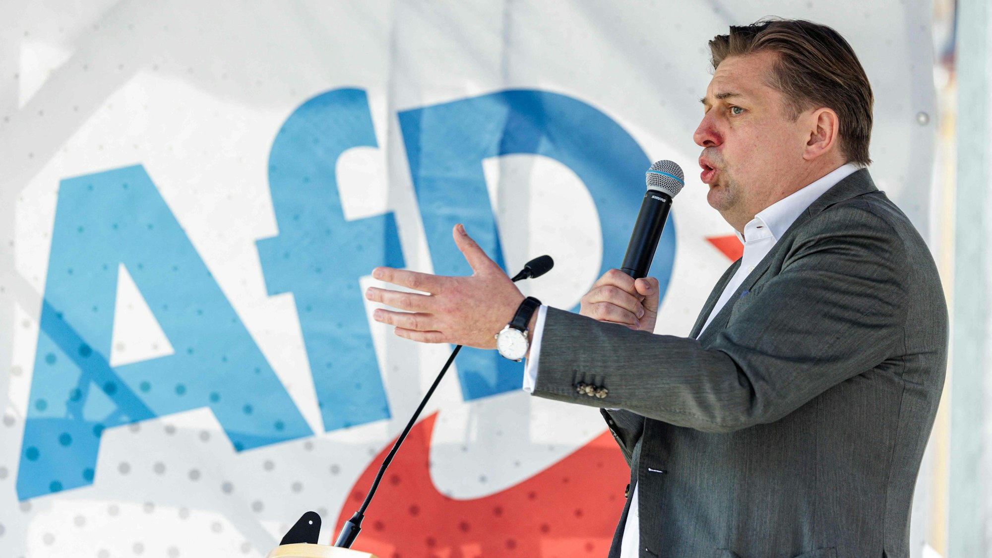 Maximilian Krah auf einer Bühne der Kundgebung in Dresden. Er hält ein Mikrofon an seinen Mund und gestikuliert mit der linken Hand. Im Hintergrund eine Flagge mit AFD-Logo.