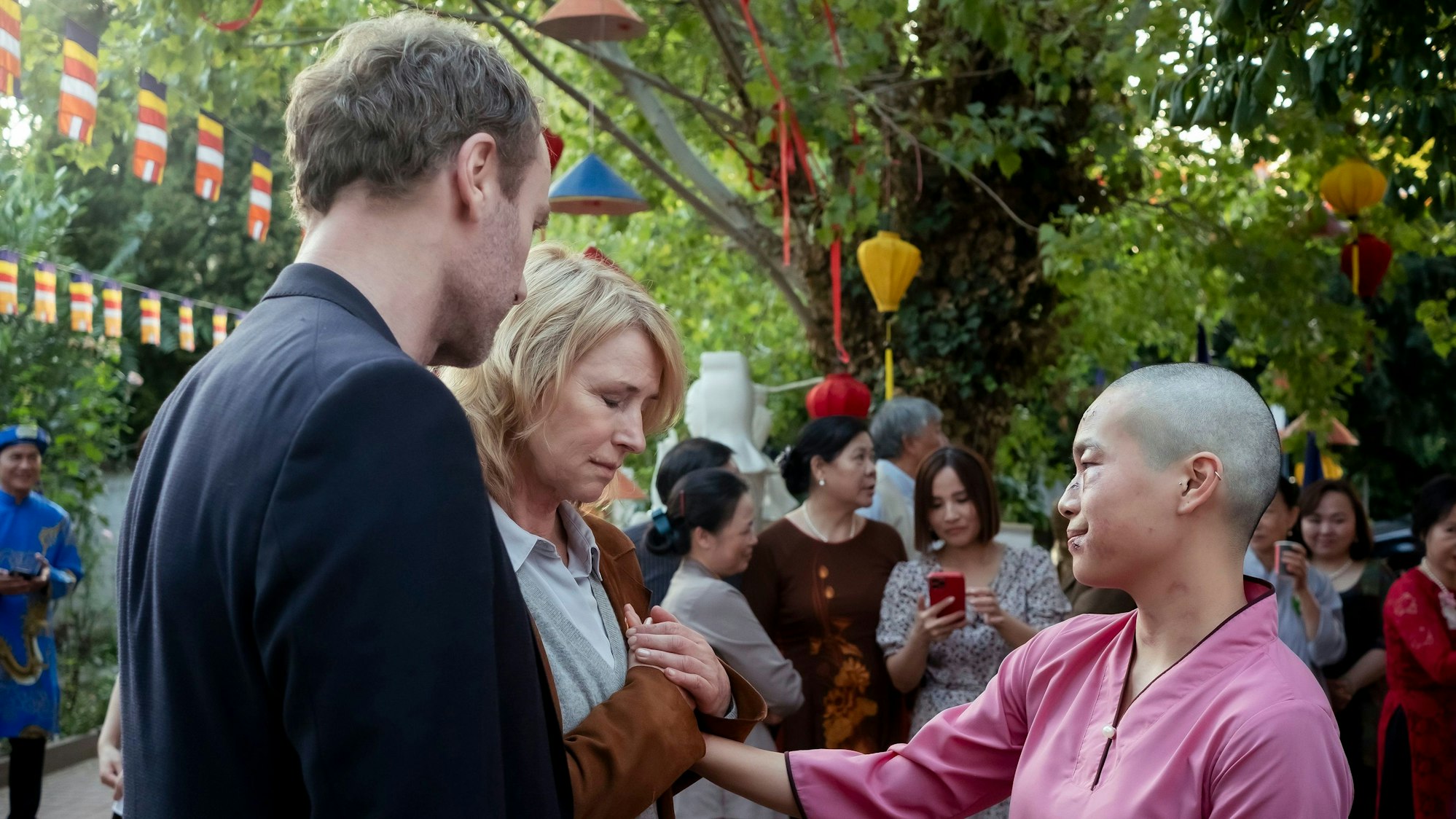 Bui Thi Vien hat den Kommissaren Blumen auf die Jacken gesteckt. Die beiden Frauen drücken einander die Hände. Kommissarin Bonard hat die Augen geschlossen. Karow steht mit dem Rücken zur Kamera und schaut auf die Geste der beiden Frauen.