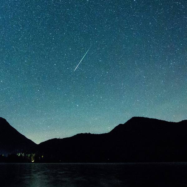 Eine Sternschnuppe leuchtet nachts neben der Milchstraße am Himmel über dem Walchensee in Bayern.