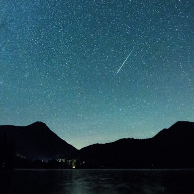 Eine Sternschnuppe leuchtet nachts neben der Milchstraße am Himmel über dem Walchensee in Bayern.
