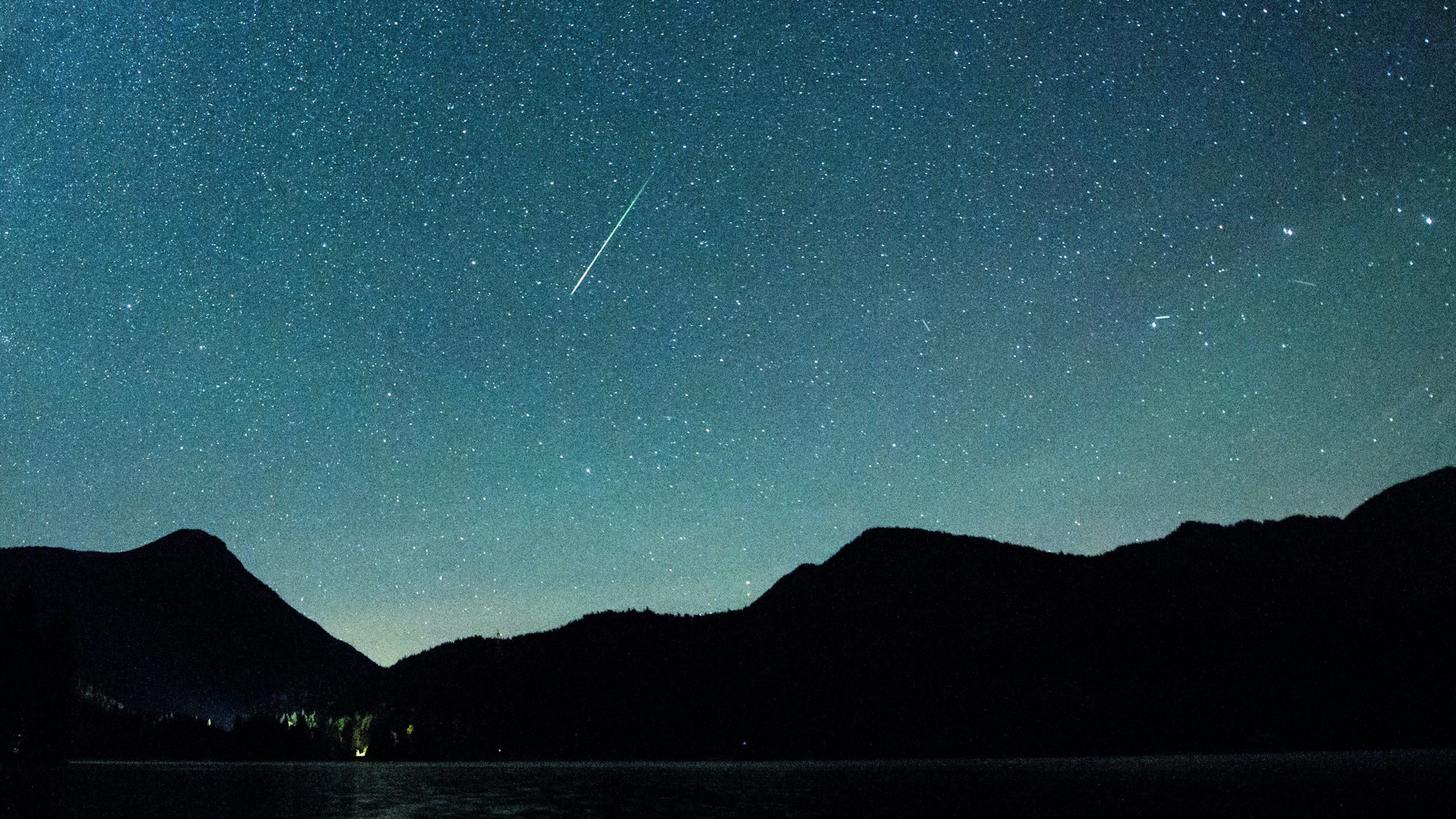 Eine Sternschnuppe leuchtet nachts neben der Milchstraße am Himmel über dem Walchensee in Bayern.