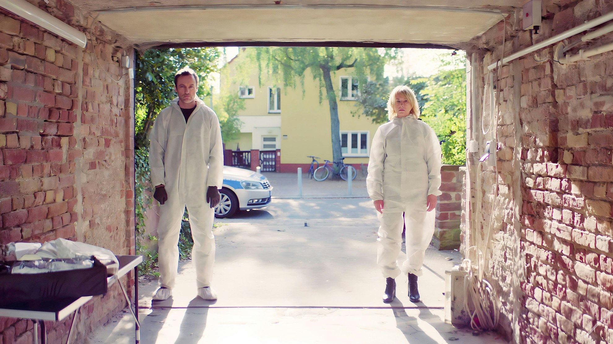 Kommissar Robert Karow und seine Kollegin Susanne Bonard stehen in weißer Schutzkleidung gebannt vor dem Kellereingang. Hinter ihnen liegt das ruhige Wohnviertel in Berlin-Lichtenberg.