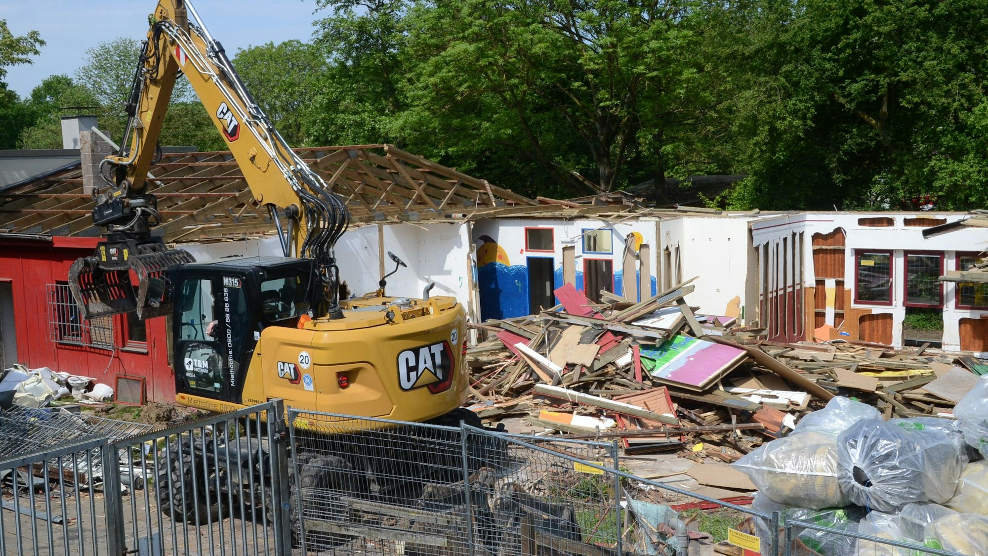 Ein Bagger steht auf einem Abrissgelände. Außenwände und ein Teil des Dachstuhls sind noch erkennbar, große Mengen Altholz liegen auf dem Boden.