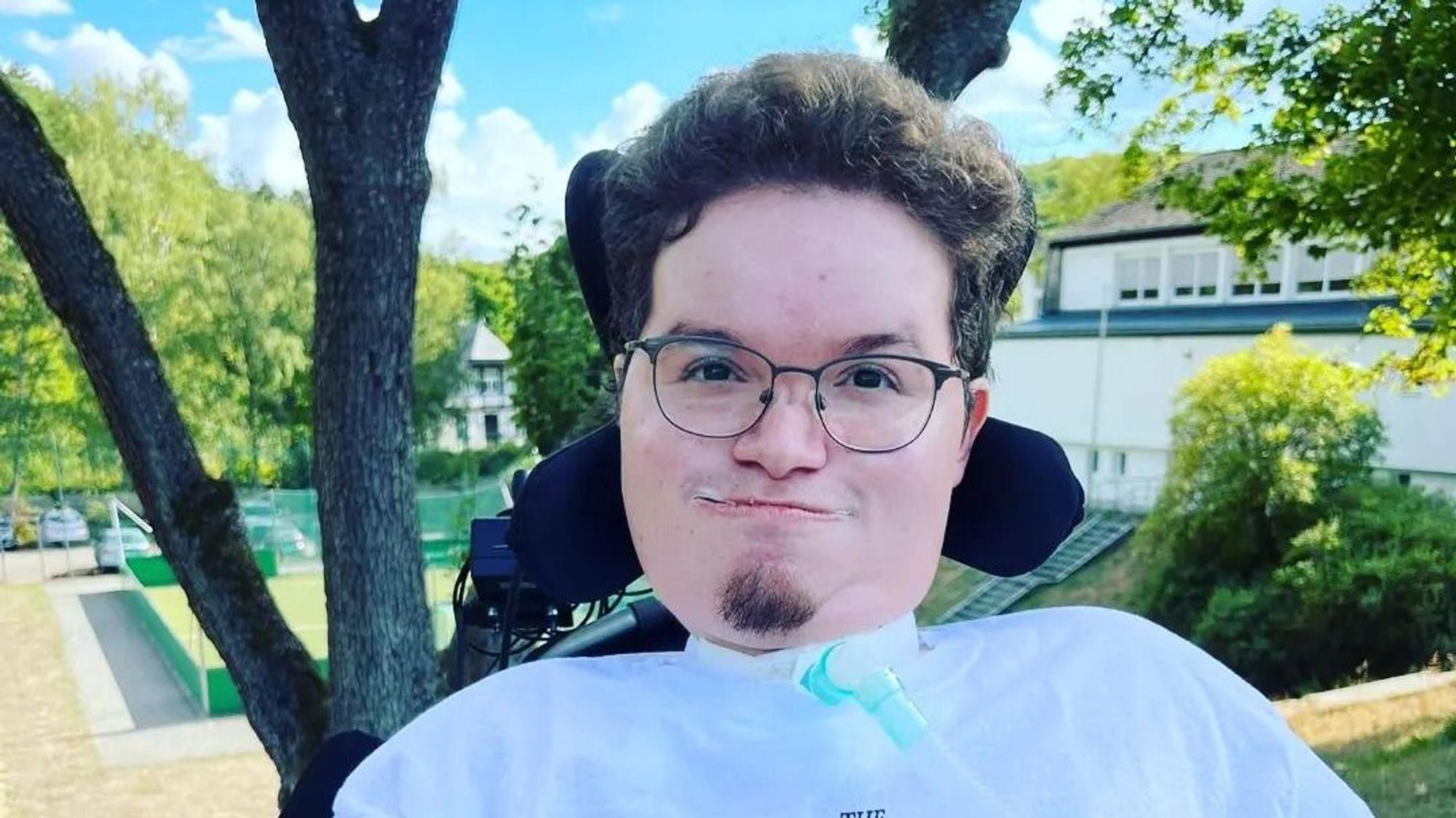 Ein junger Mann mit Brille und Kinnbart sitzt in einem Rollstuhl.