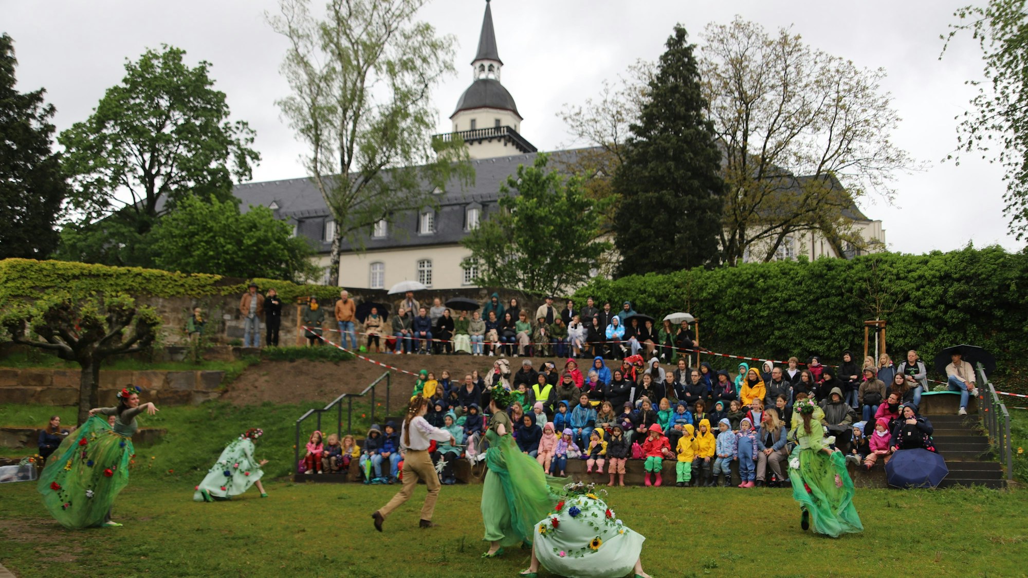 Die Feen tanzen im Johannisgarten vor ihrem Publikum, mit der ehemaligen Abtei im Hintergrund.