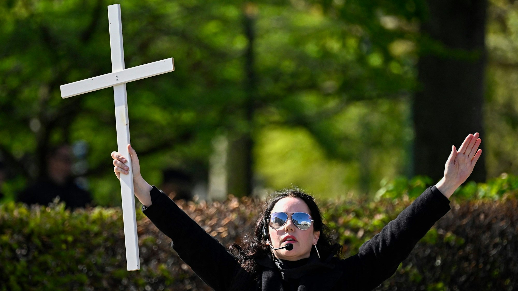 Eine Frau mit Sonnenbrille und Kopfmikrofon breitet die Arme weit aus. In ihrer rechten Hand hält sie ein Kreuz.
