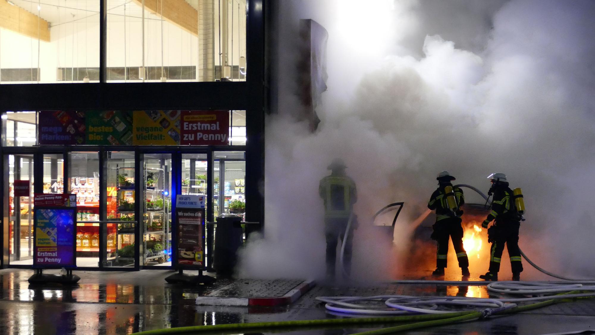 Feuerwehrleute löschen ein brennendes Auto nebem dem Eingang eines Supermarkts.