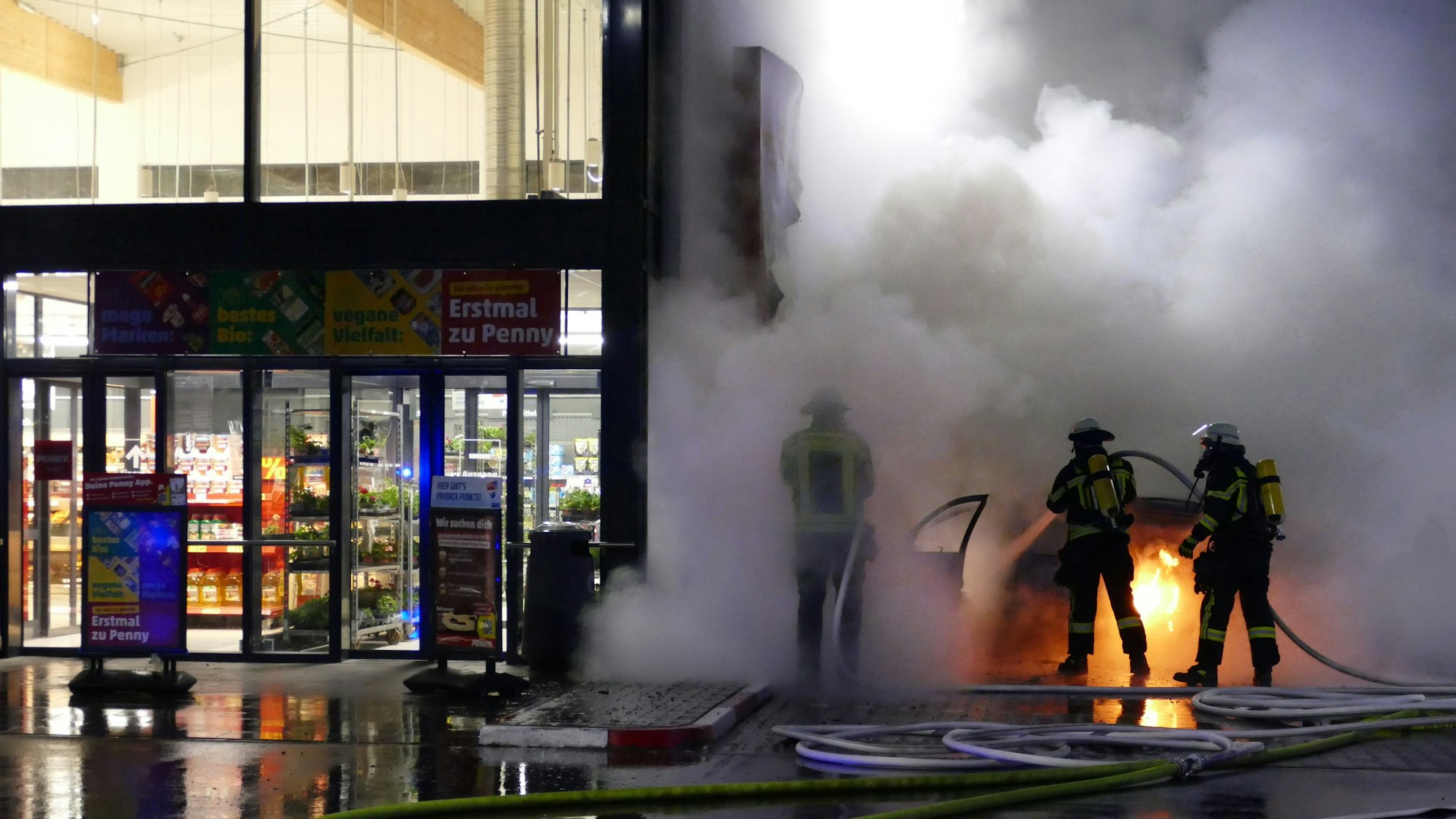 Feuerwehrleute löschen ein brennendes Auto nebem dem Eingang eines Supermarkts.
