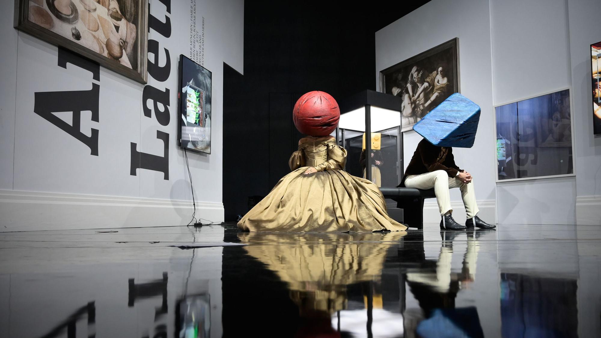 Frau und Mann sitzen in Biedermeier-Kostümen auf einer Bank in der Kulisse von Claudia Bossards „Baracke“-Inszenierung am Deutschen Theater Berlin. Hinten steht zwischen ihnen ihr Kind in einem Glaskasten. Die Frau trägt einen roten Ball über ihren Kopf, der Mann ein blaues Dreieck, das Kind einen gelben Legostein-Quader.